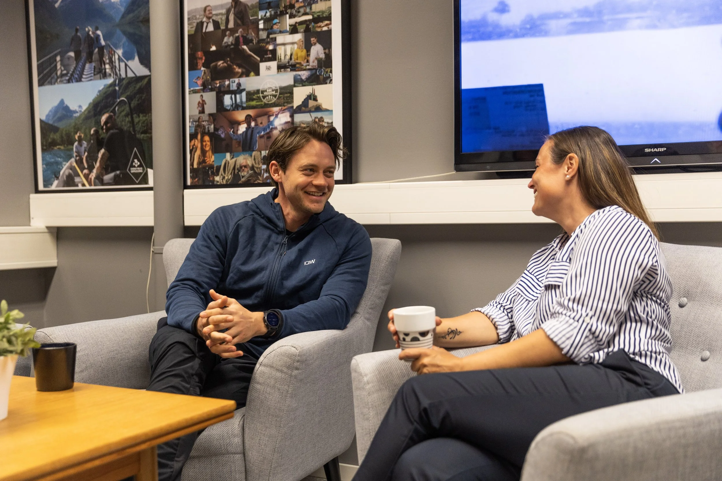 Two people, a man and a woman, are sitting and smiling at each other in a casual meeting room. The woman is holding a coffee mug, and the man is wearing a navy hoodie. Behind them are posters with photos of outdoor activities and a large TV screen on the wall.