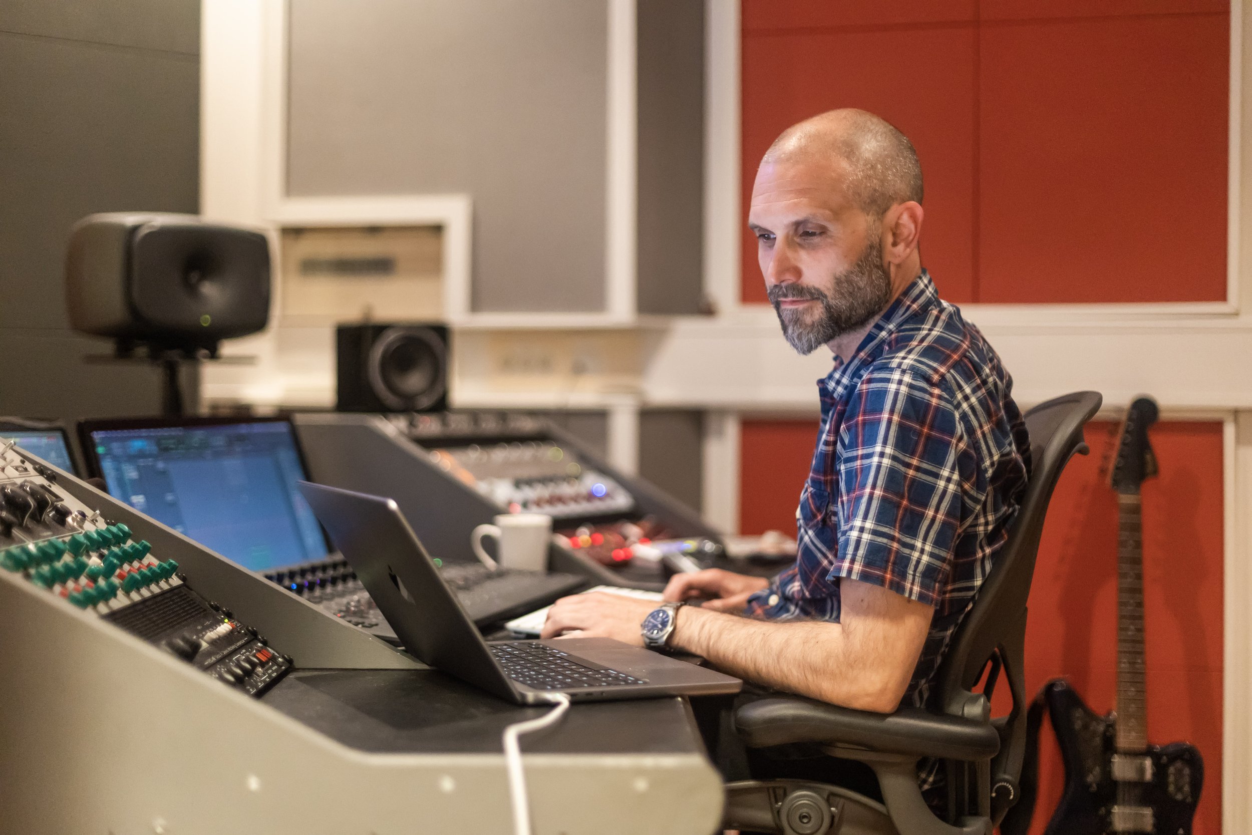 A man with a beard wearing a plaid shirt working at a music studio with audio equipment and speakers.