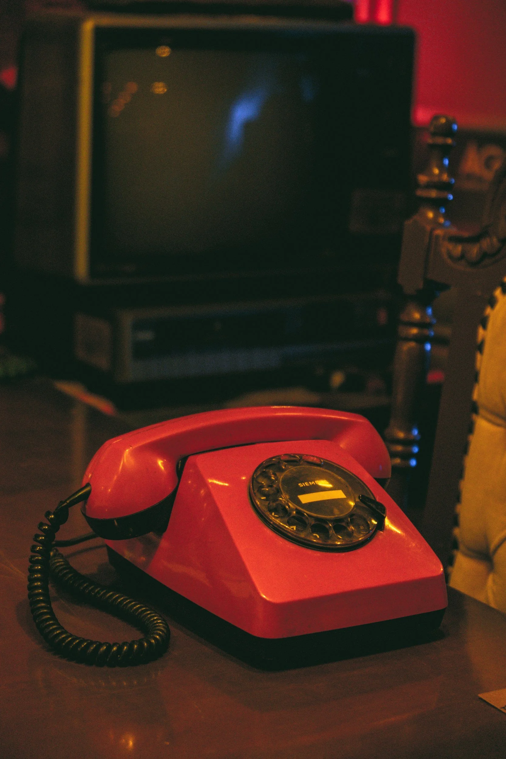 A red vintage rotary telephone placed on a dark table. In the background, there is an old television or monitor turned off, with warm lighting and a red wall.
