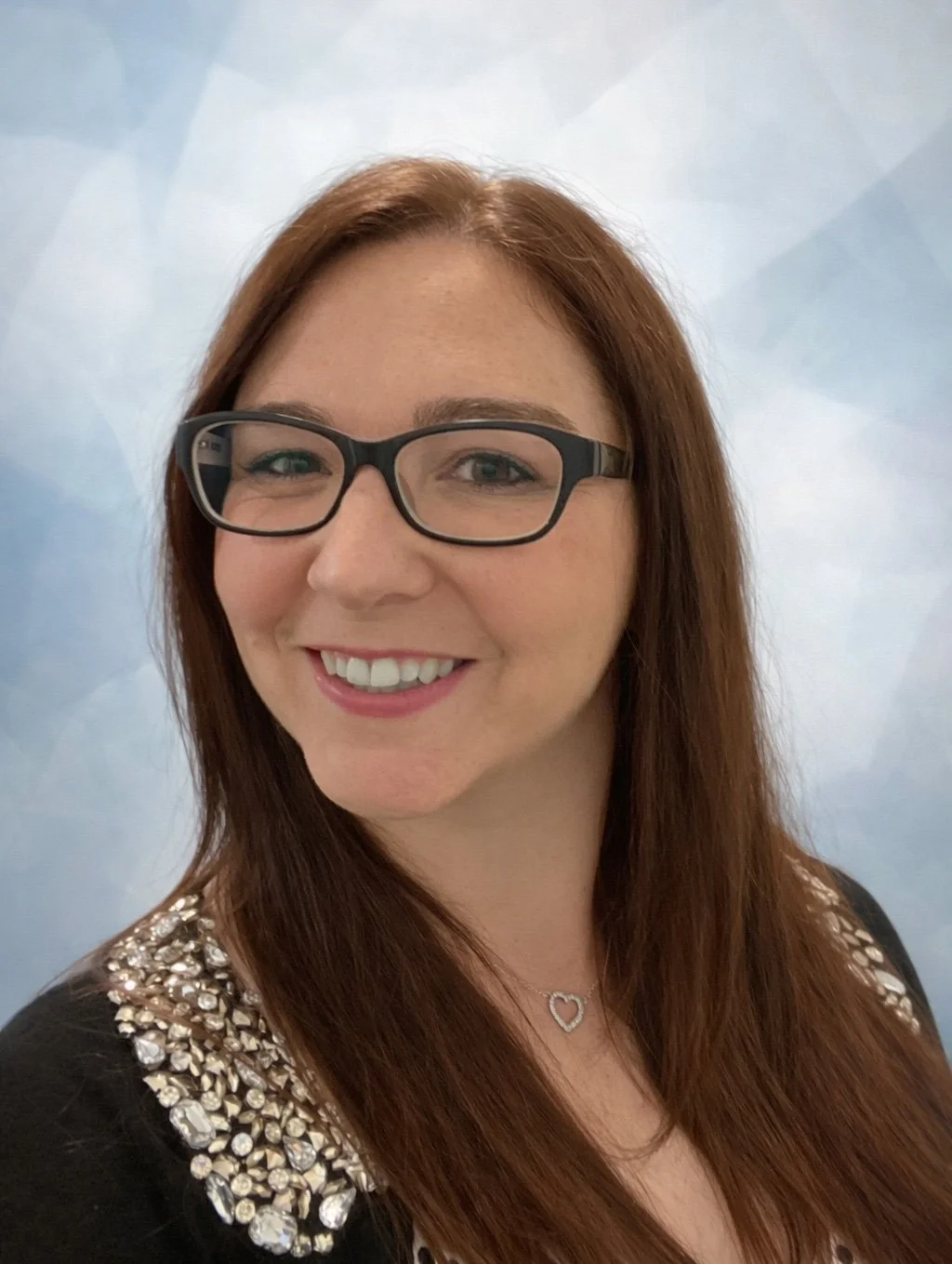A woman with long red hair and glasses smiling against a soft blue background.