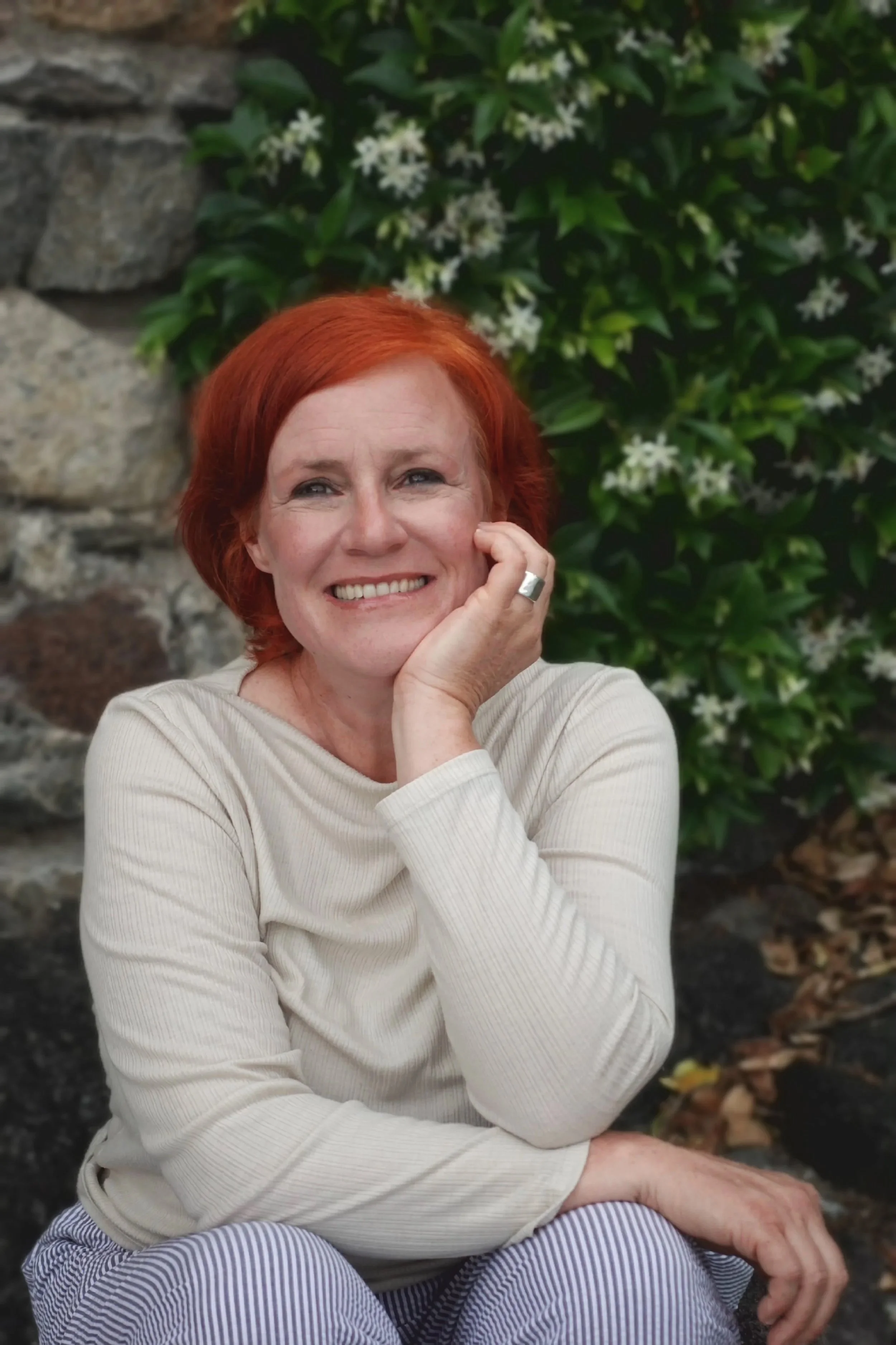 A woman with red hair smiling and resting her head on her hand outdoors, surrounded by green foliage and white flowers.