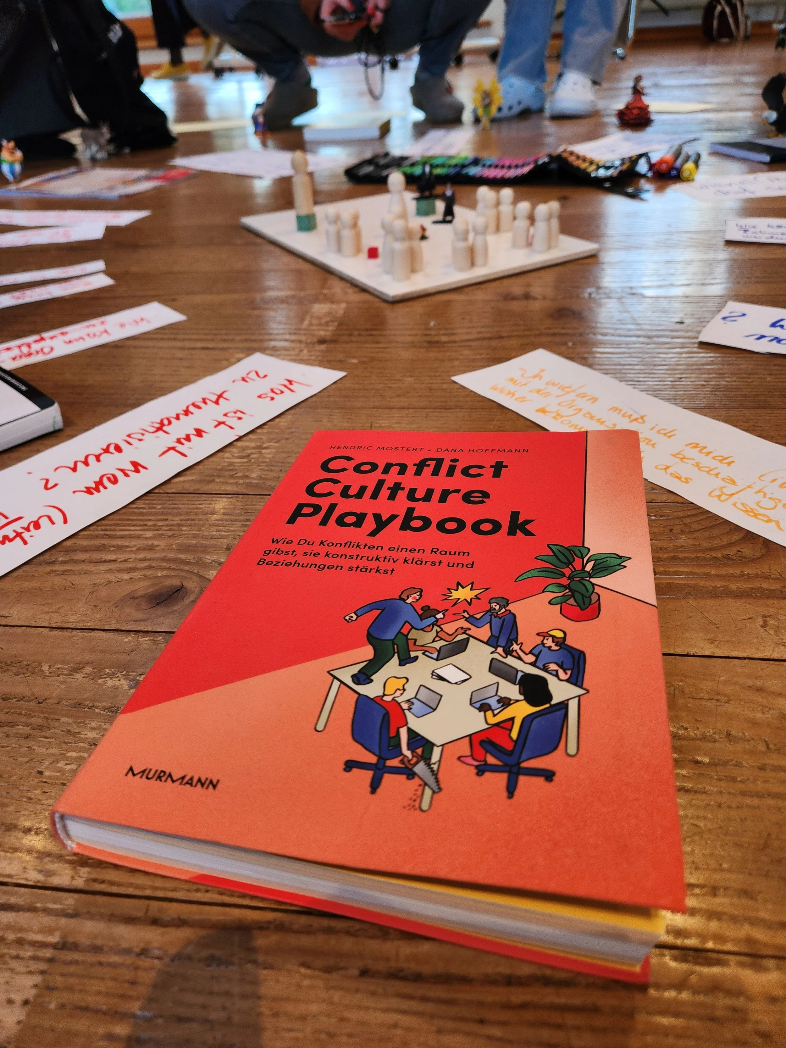 A colorful book titled "Conflict Culture Playbook" on a wooden table, featuring an illustration of people around a table engaged in a discussion. In the background, there are handwritten notes, markers, and a game setup with wooden figures on a board.