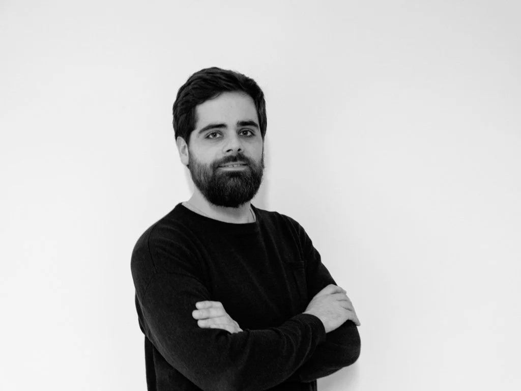 Hombre con barba y cabello oscuro, con brazos cruzados, usando una camiseta negra, de pie frente a una pared blanca.