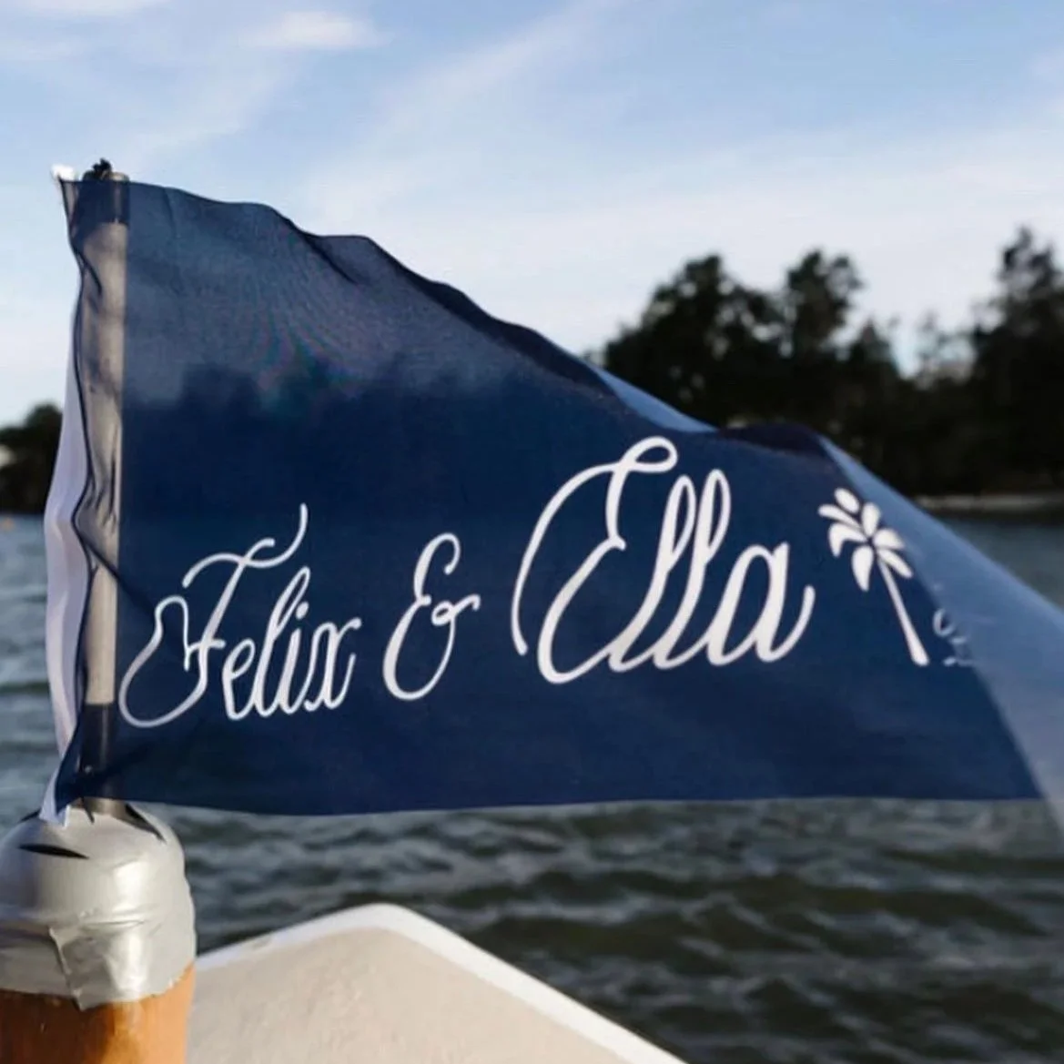 Close-up of a boat flag with the names Felix & Ella written in white cursive on a blue background, mounted on a white pole, with water and trees in the background.