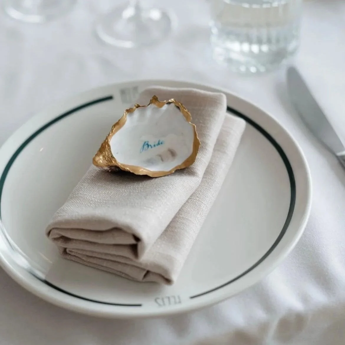 A white plate with a gold-edged oyster shell resting on a folded beige napkin, with a glass of water and cutlery in the background on a white table.