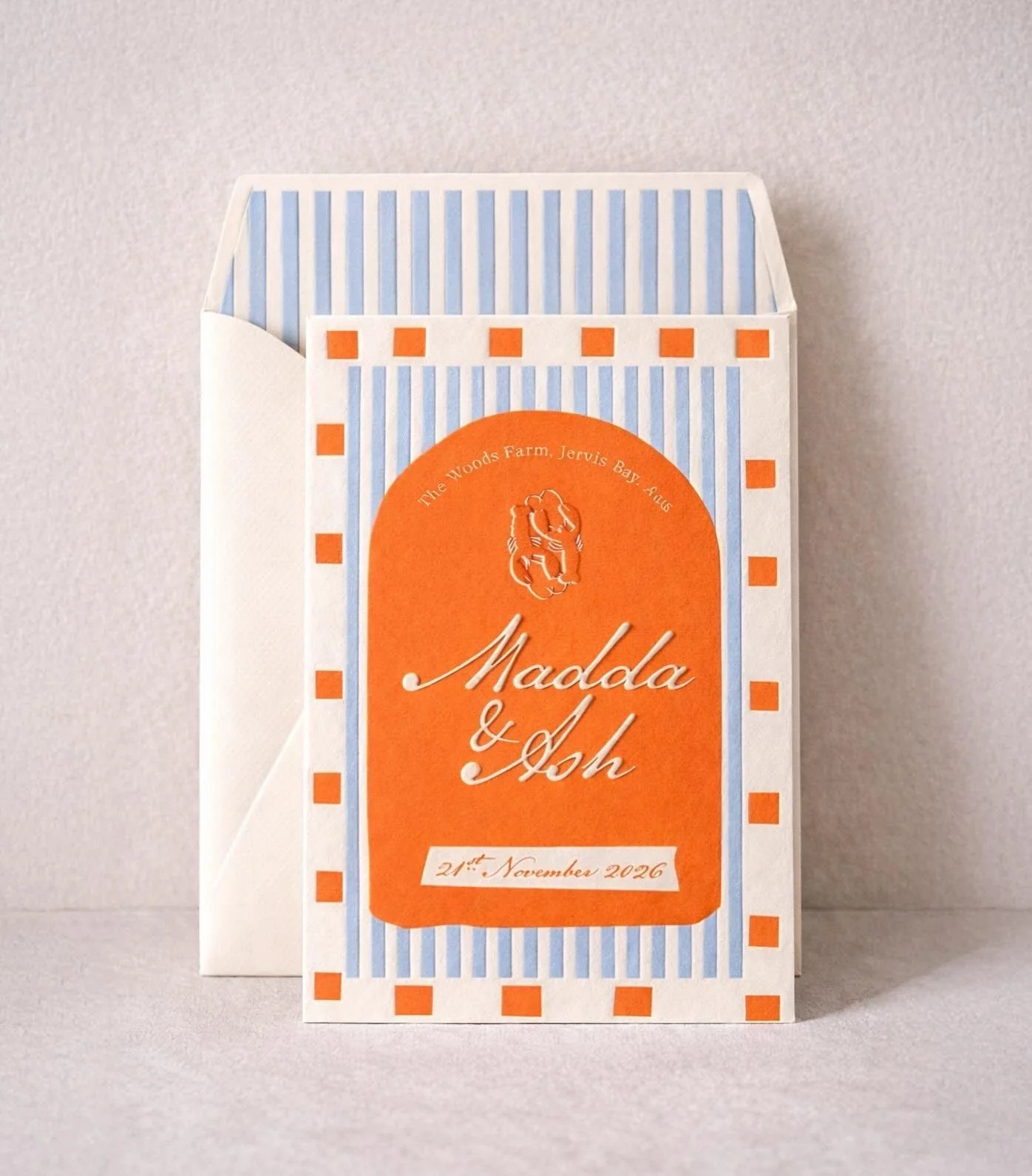 A wedding invitation with orange and blue design elements, displaying the names Madola and Ash, and the wedding date, 21st November 2026, on a gray surface.