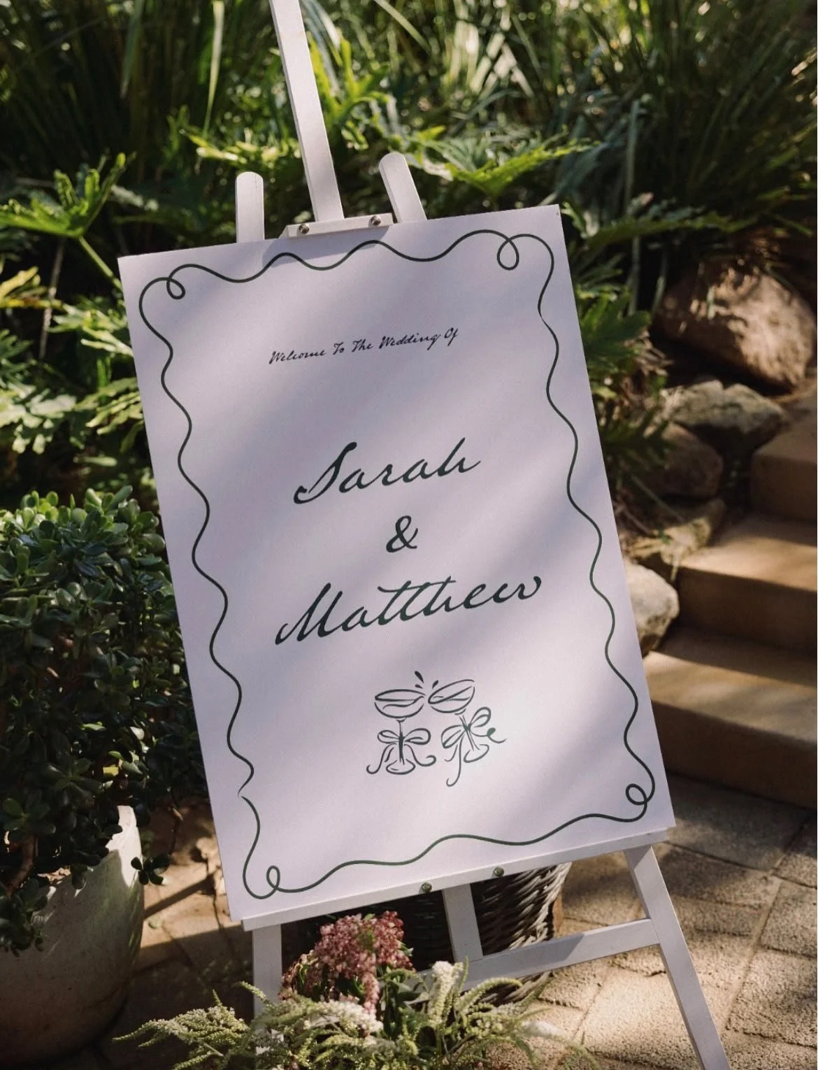 Wedding welcome sign with the names Sarah and Matthew, decorated with floral illustrations, placed outdoors among greenery and rocks.