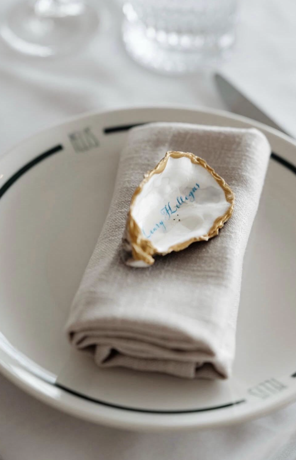 A seashell with gold edges and blue writing inside, placed on a folded beige napkin on a white plate, with a blurred glass in the background.