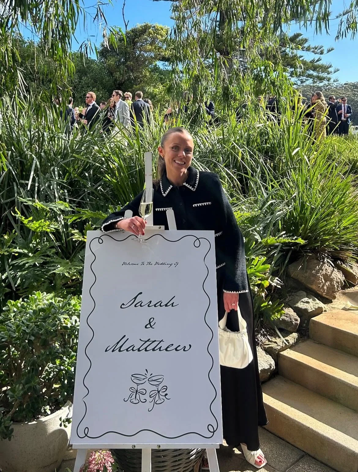 A woman at an outdoor wedding reception holding a glass of champagne and a bag, standing beside a sign that reads 'Welcome to the wedding of Sarah & Matthew' with decorative illustrations, greenery, and a crowd of guests in the background.