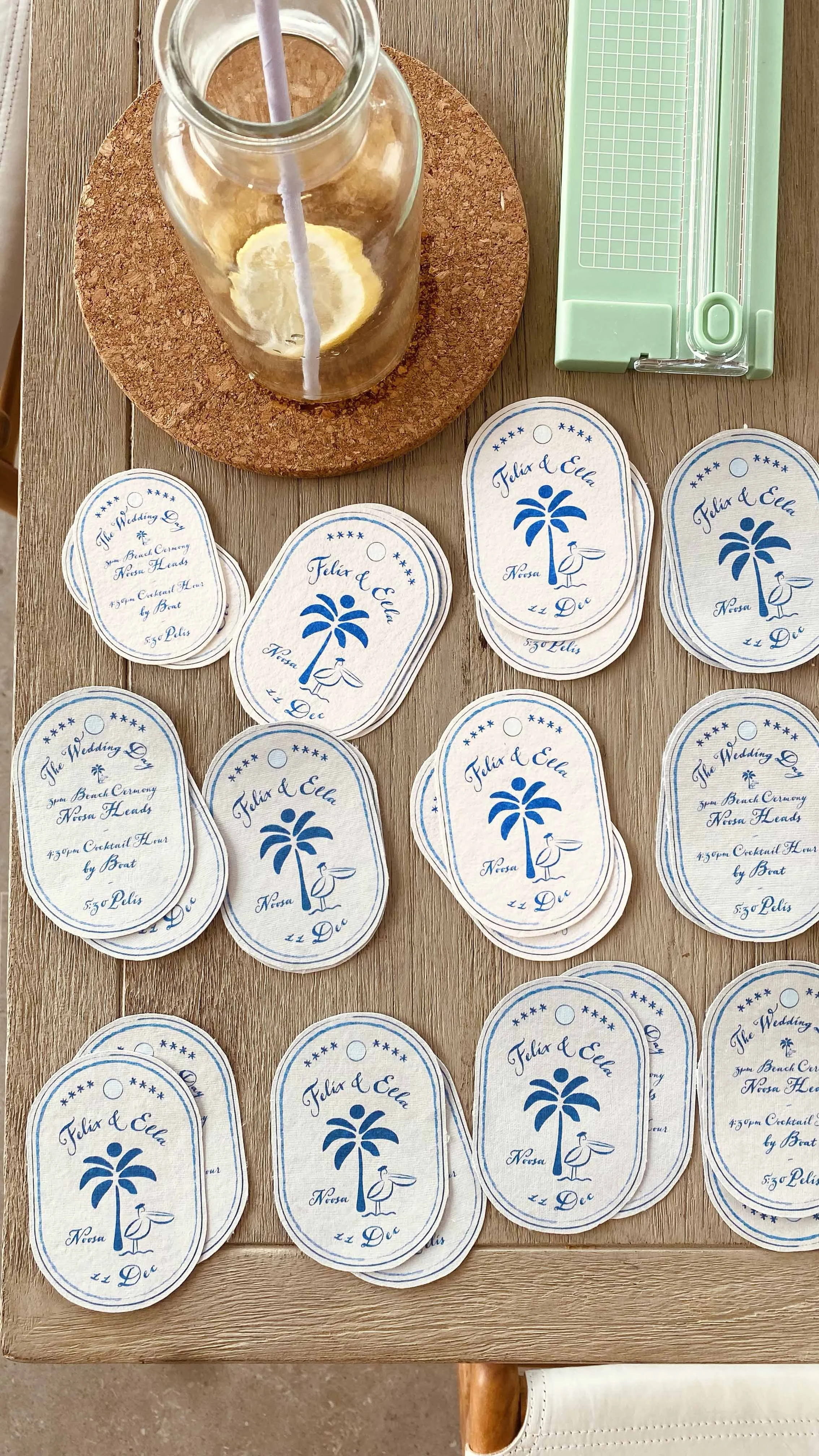 Wedding event place cards with blue palm trees and a pelican, labeled with the names Felix and Ella, date December 11th, time 4:30 PM, location Noosa. A glass bottle with lemon slices and a straw is on a cork placemat, and a green paper cutter is beside it on a wooden table.