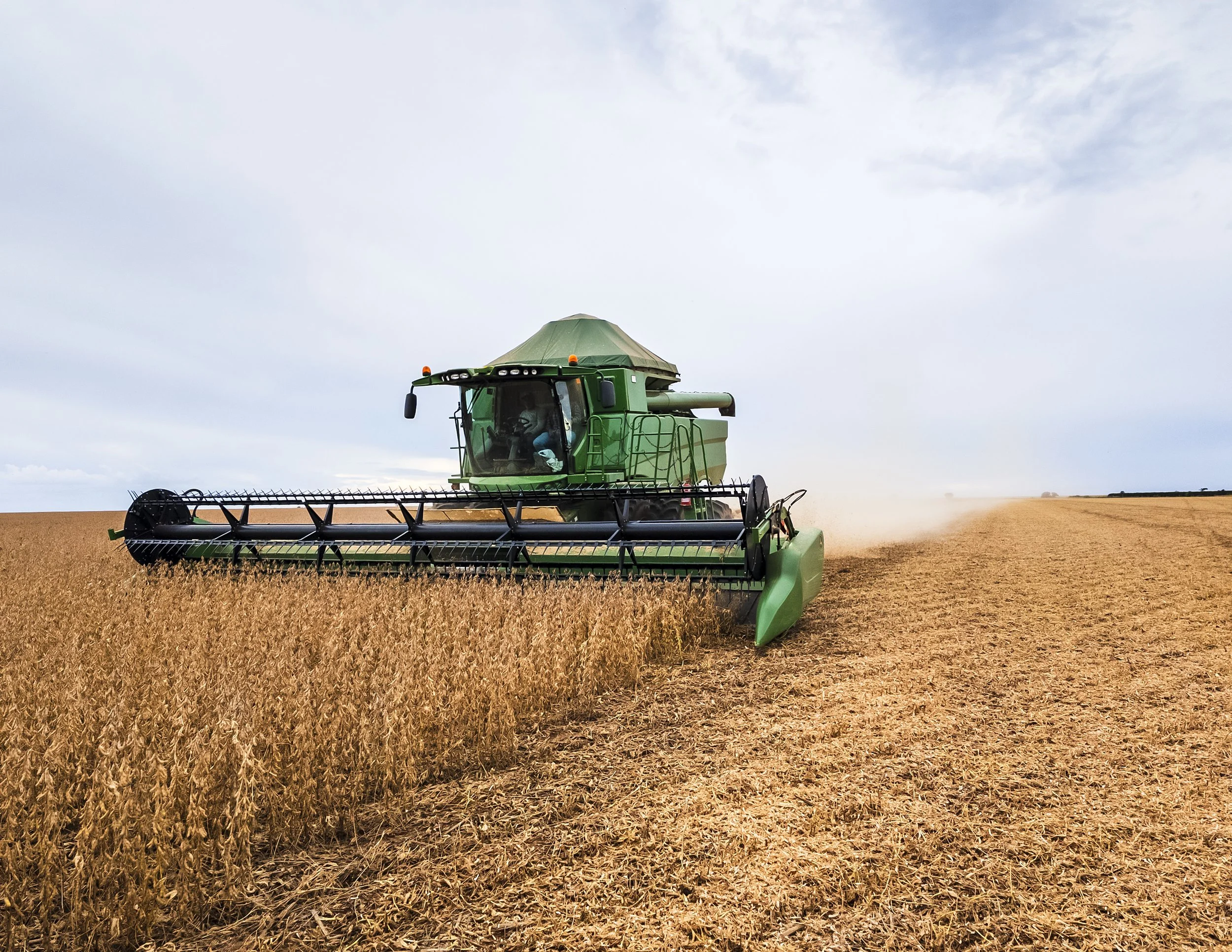 A green combine harvester operating in a vast wheat field under cloudy sky, harvesting crops.