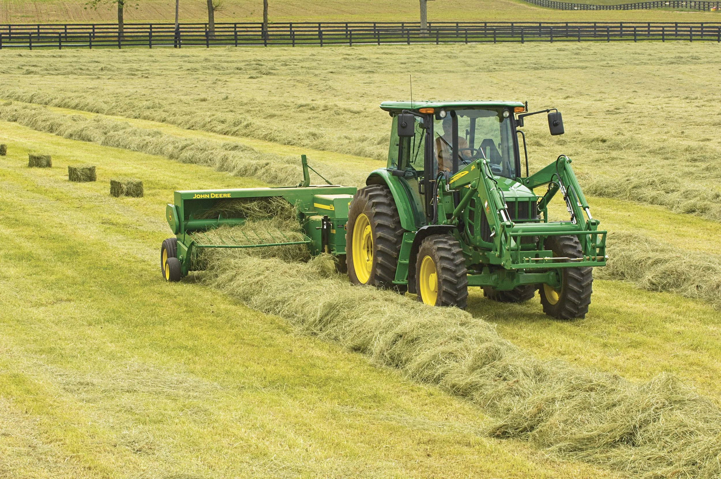 A green John Deere tractor harvesting hay in a large pasture. Hay bales are scattered around the field.
