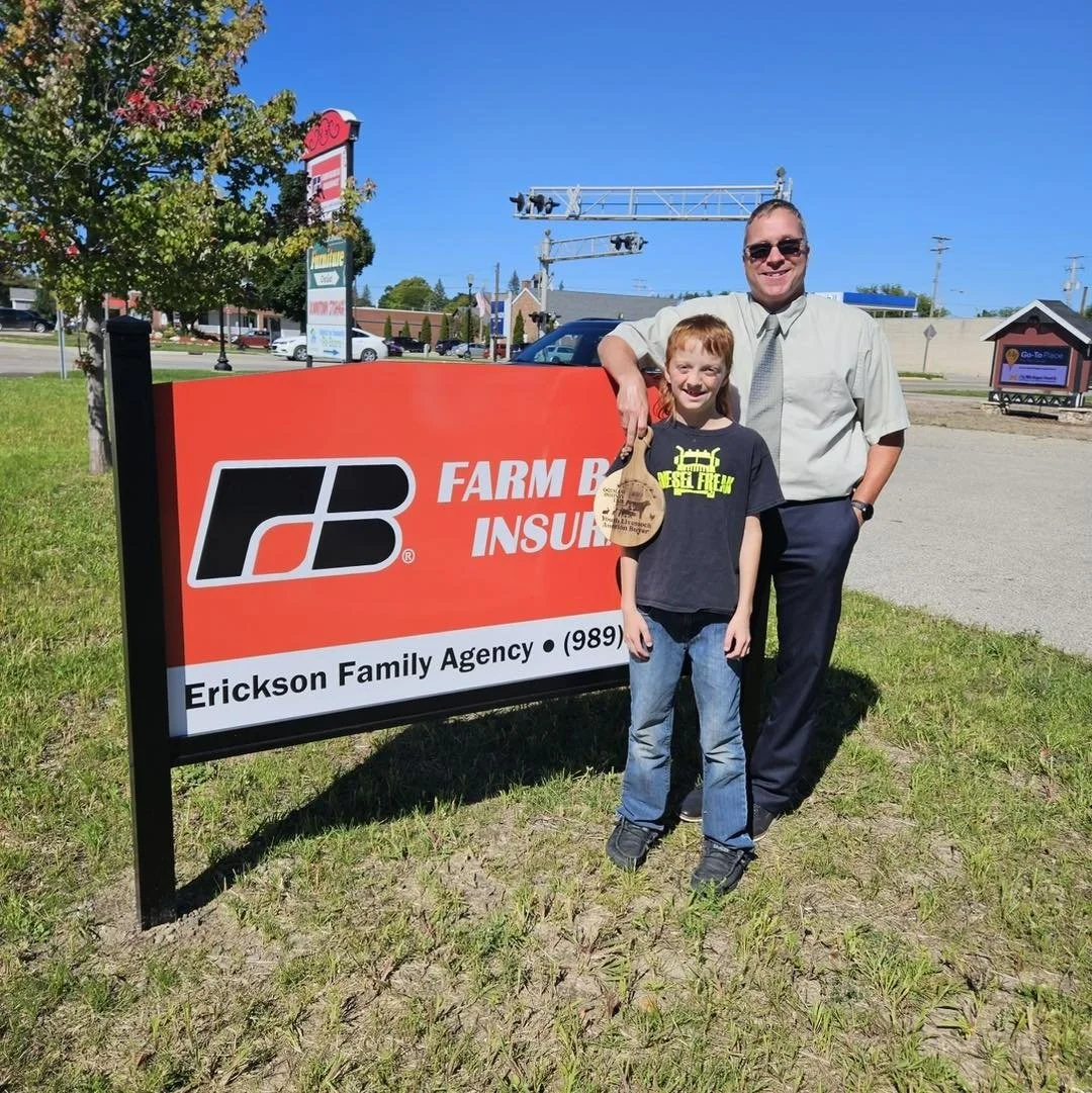A man and a young girl standing next to a Ford Farm Bureau Insurance sign on a sunny day. The man is wearing sunglasses, a light shirt, and dark pants, with his arm around the girl. The girl has red hair, a black t-shirt, and blue jeans, holding a paper bag. In the background, there are trees, a parking lot with cars, and a train track with signals.