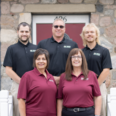 Group of five smiling people standing outdoors in front of a stone building with a red door, three men wearing black polo shirts and two women wearing matching maroon shirts.
