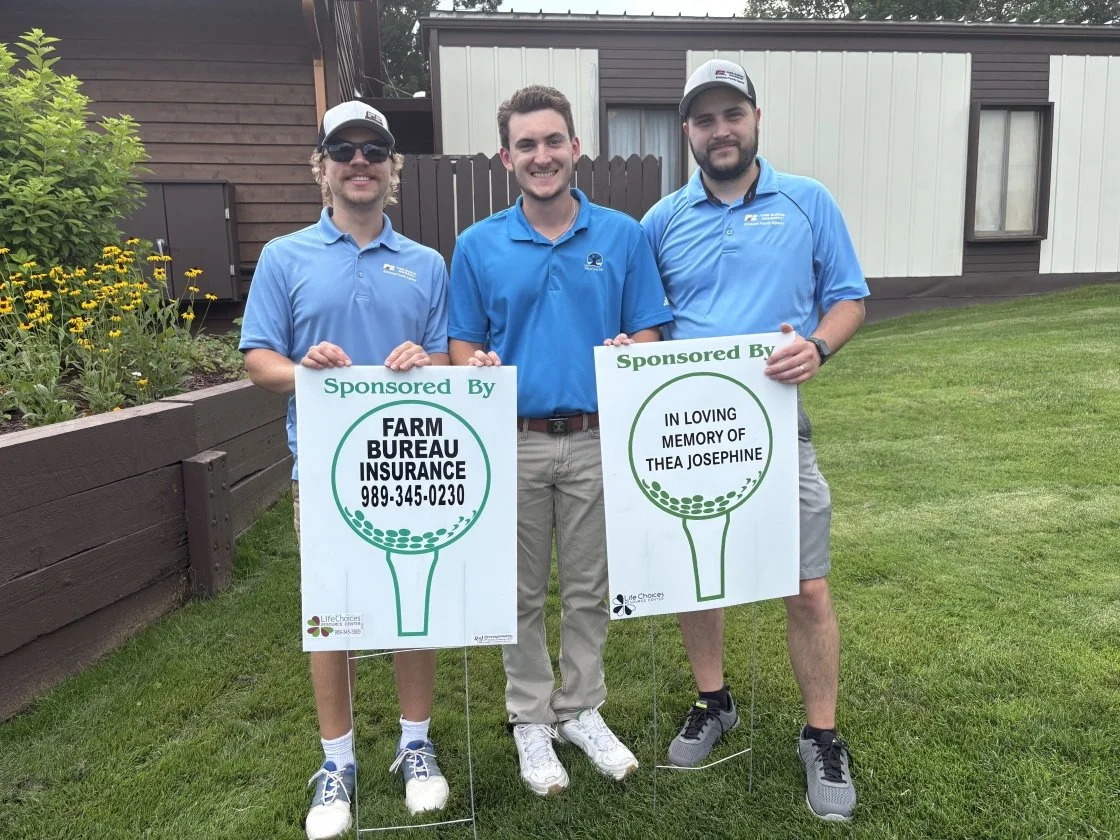 Three young men standing outdoors on grass, holding signs, smiling. Two wear sunglasses and gray caps, all wear blue shirts. Background has a wooden garden bed with yellow flowers and a small building.