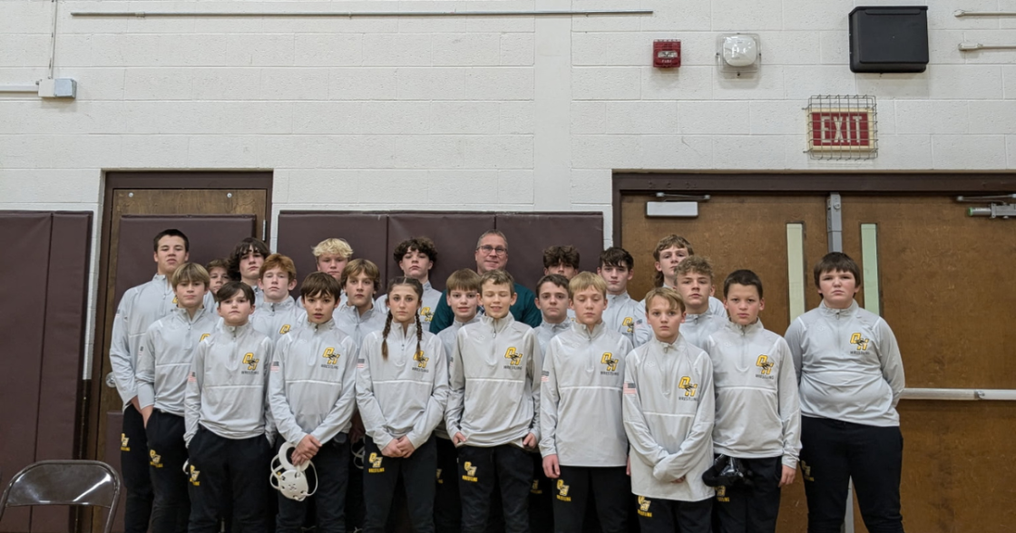 Group of young boys and one adult man standing in gymnasium, wearing matching gray athletic jackets with a logo, some holding a white sports helmet.