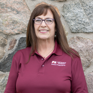 A woman with shoulder-length brown hair and glasses smiling in front of a stone wall, wearing a maroon polo shirt with a logo on the left side.