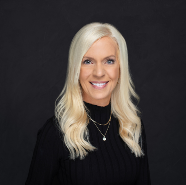 A woman with long blonde hair smiling, wearing a black top and gold jewelry, against a dark background.