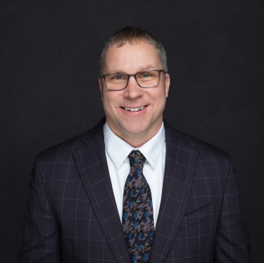 Portrait of a smiling middle-aged man with glasses in a dark plaid suit, white shirt, and patterned tie, against a dark background.