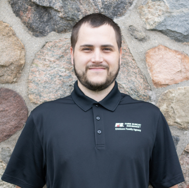 A man with short dark hair and a beard smiling, wearing a navy blue polo shirt with a FFA logo, standing outdoors in front of a stone wall.