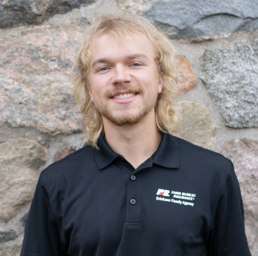 Portrait of a young man with blond hair and a beard smiling, wearing a black polo shirt with a logo on the left side, standing outdoors against a stone wall background.