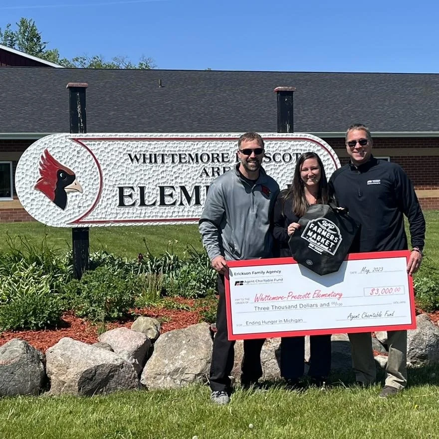Three people holding a large check for $3,000 in front of a sign for Whittemore Prescott Elementary School, with a backpack and a smiling woman, on a sunny day.