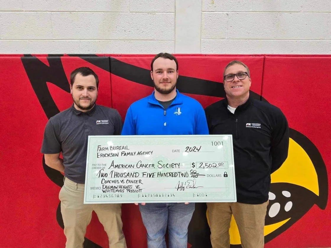 Three men stand in front of a red backdrop with a yellow and black logo, holding a large check made out to the American Cancer Society for $2,502. The man in the middle is wearing a blue jacket, the man on the left is in a gray polo, and the man on the right is in a black jacket with glasses.