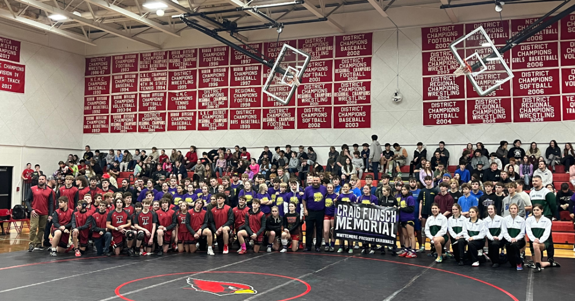 Group of wrestlers, coaches, and fans posing for a team photo in a gymnasium, with banners on the wall and a wrestling mat in the foreground.