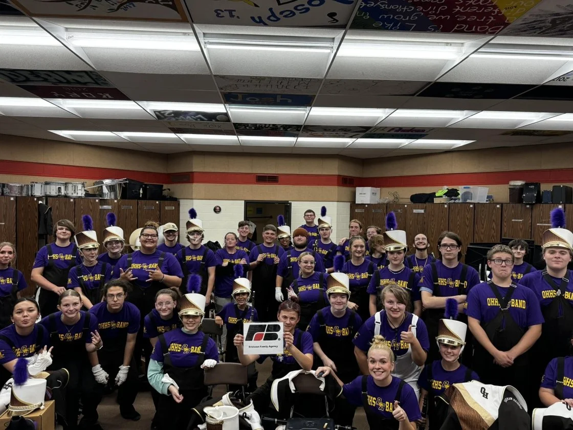 A group of young band members wearing purple shirts and marching band hats, posing inside a school locker room with lockers in the background.