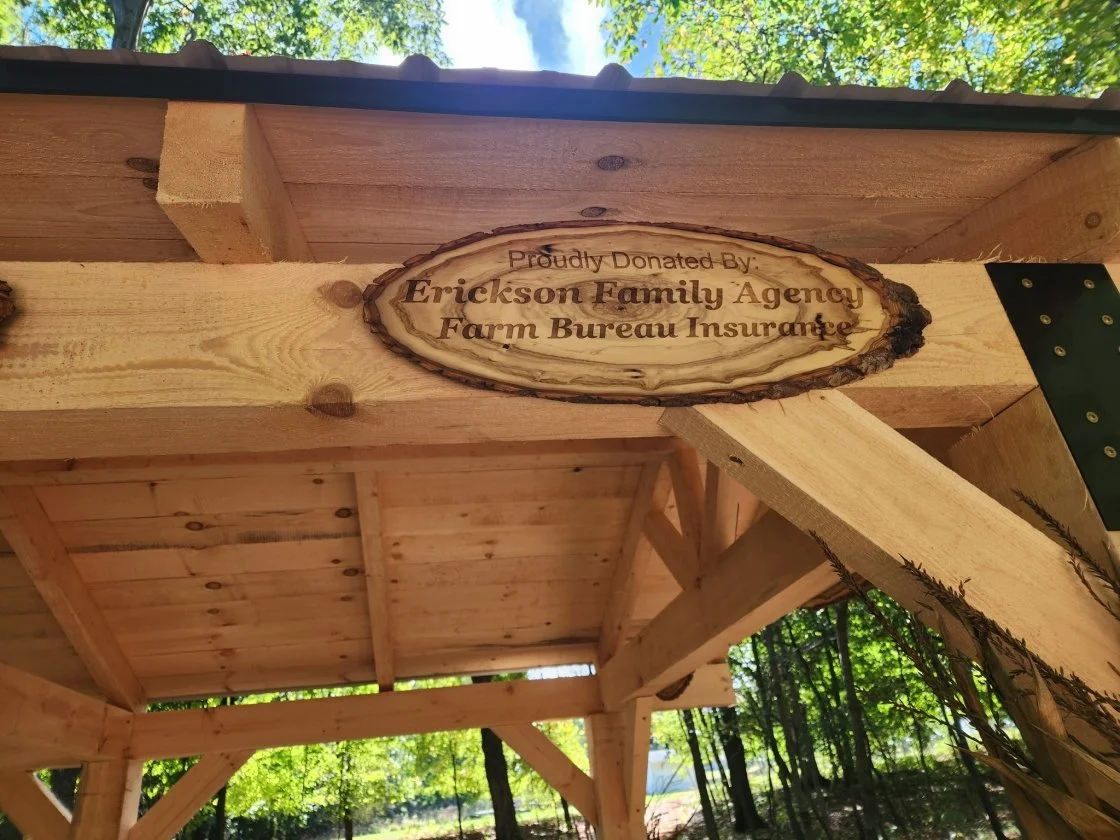 Wooden sign inside a wooden shelter with trees in the background, reading 'Proudly Donated By: Erickson Family Agency Farm Bureau Insurance'.