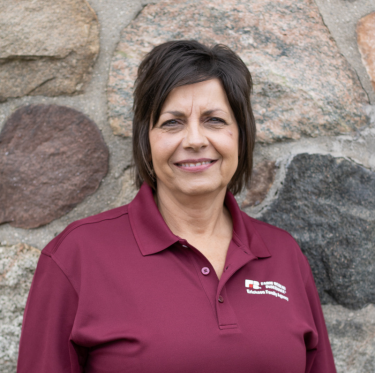Woman with short dark hair smiling, wearing a maroon polo shirt with a logo, standing in front of a stone wall.