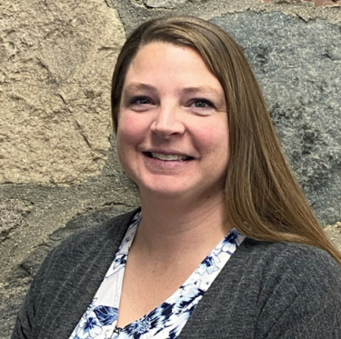 A woman with long light brown hair, smiling, wearing a gray blazer over a white and blue patterned shirt, standing in front of a stone wall.
