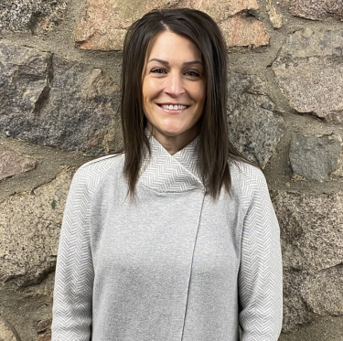 A woman with dark brown hair, smiling, standing in front of a stone wall, wearing a light gray jacket.
