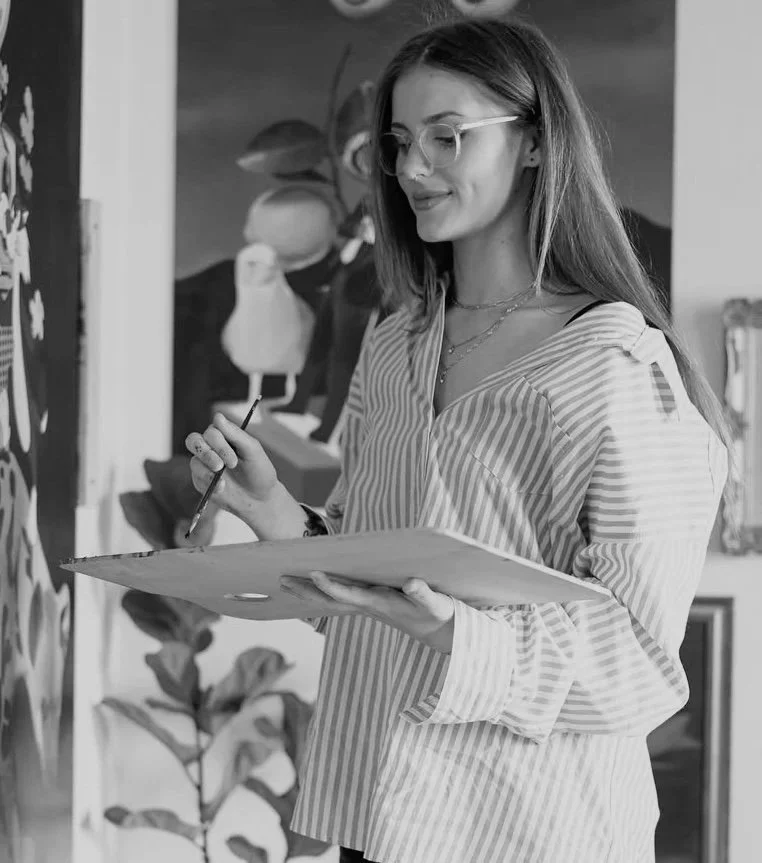 A woman wearing glasses and a striped shirt is holding a paint palette and brush, standing in an art studio with plants and artwork in the background.