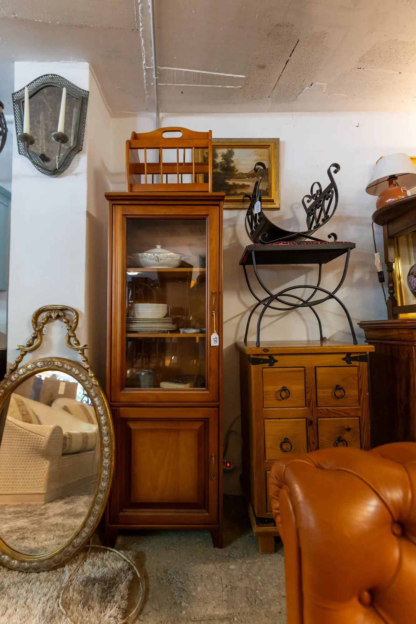 A collection of vintage furniture and decor including a wooden display cabinet, a carved wooden desk, a leather armchair, an ornate mirror, a wall lamp, and a framed landscape painting. Aymerick Rondeau Furniture Emporium Alhaurin second hand furnitu