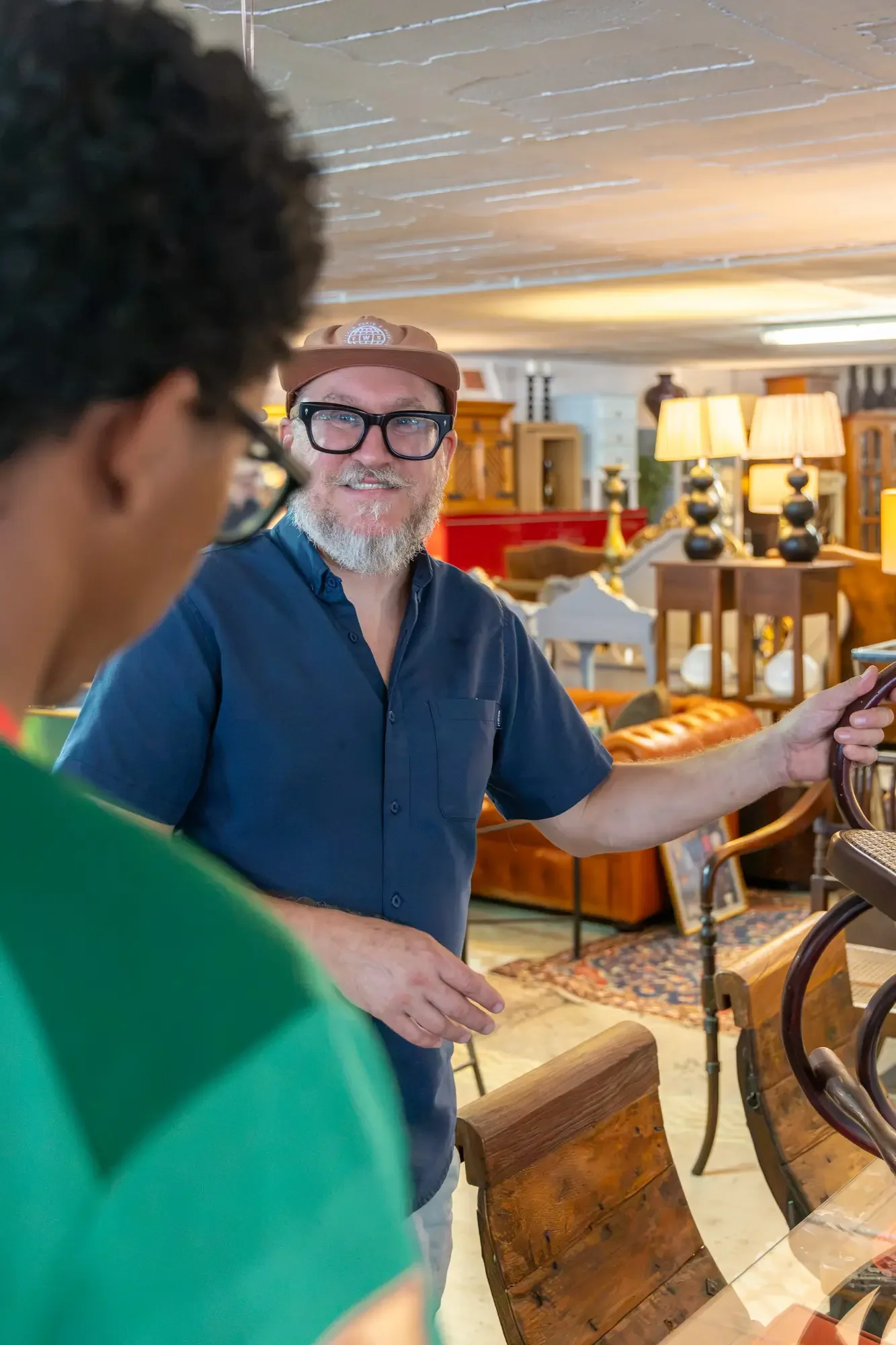 A man with glasses and a beard, wearing a tan cap, is smiling while looking at his phone. He is inside a store with wooden furniture and lamps in the background. Part of a person in a green shirt is visible in the foreground. Aymerick Rondeau Furnitu