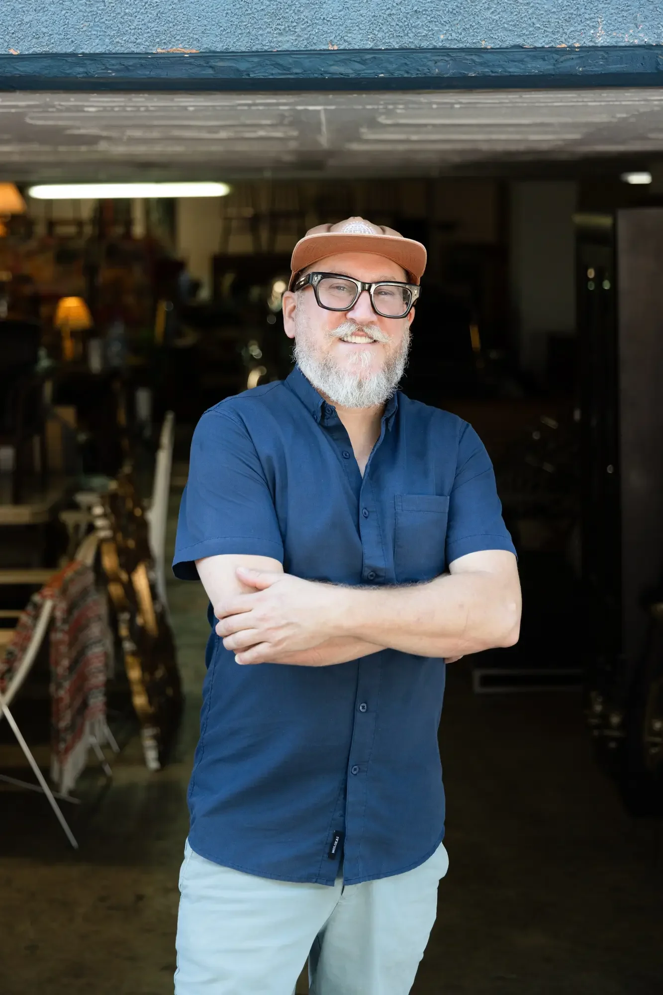 A man with glasses, a beard, and a cap smiling and crossing his arms in a cozy indoor setting. Aymerick Rondeau Furniture Emporium Alhaurin second hand furniture shop