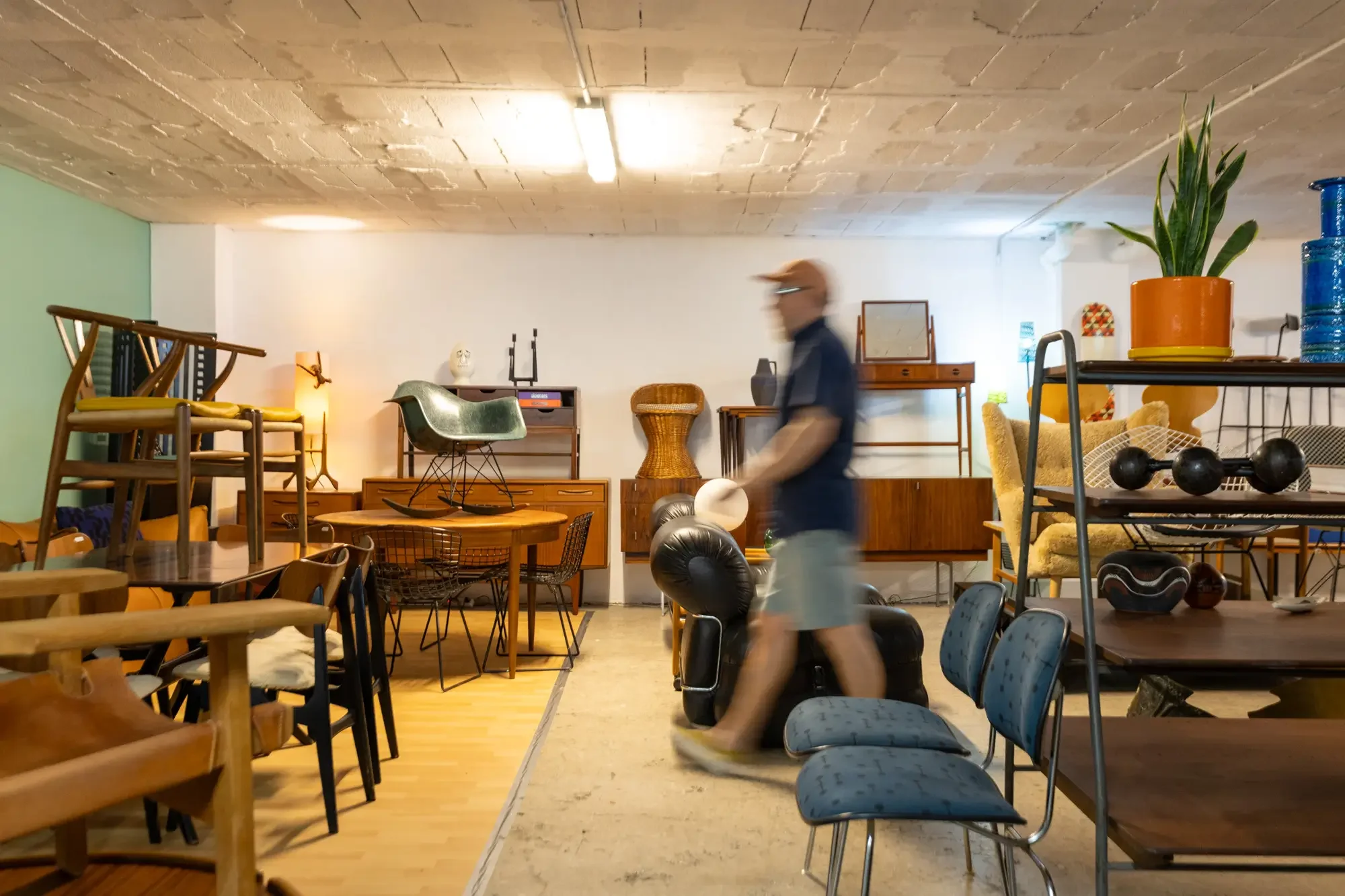 A person walking past a cluttered furniture and decor store with chairs, tables, lamps, plants, and various vintage and modern items. Aymerick Rondeau Furniture Emporium Alhaurin second hand furniture shop