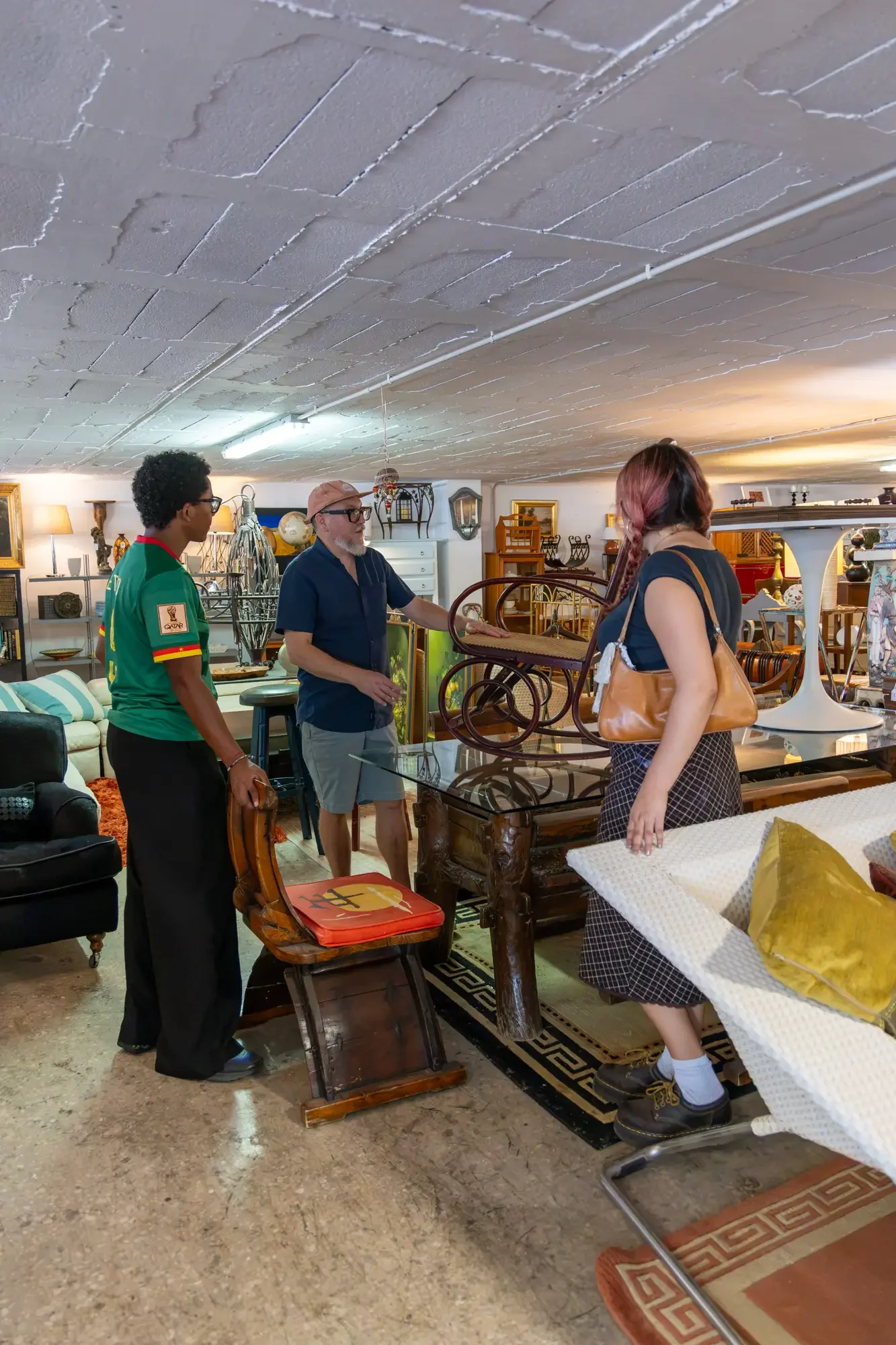 Three people standing around a wooden table in a room filled with various furniture and decorative items, with one person holding an orange bag with a black stripe, and all engaged in conversation. Aymerick Rondeau Furniture Emporium Alhaurin second