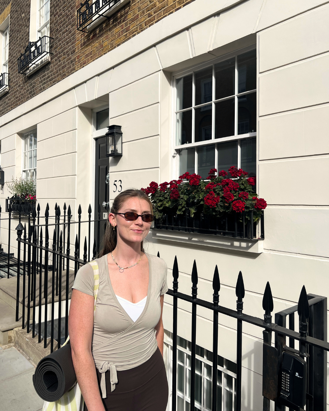 A woman with brown hair, wearing sunglasses, a beige wrap top, black pants, and a white shirt underneath, standing in front of a white building with black iron fence and red flower window box.