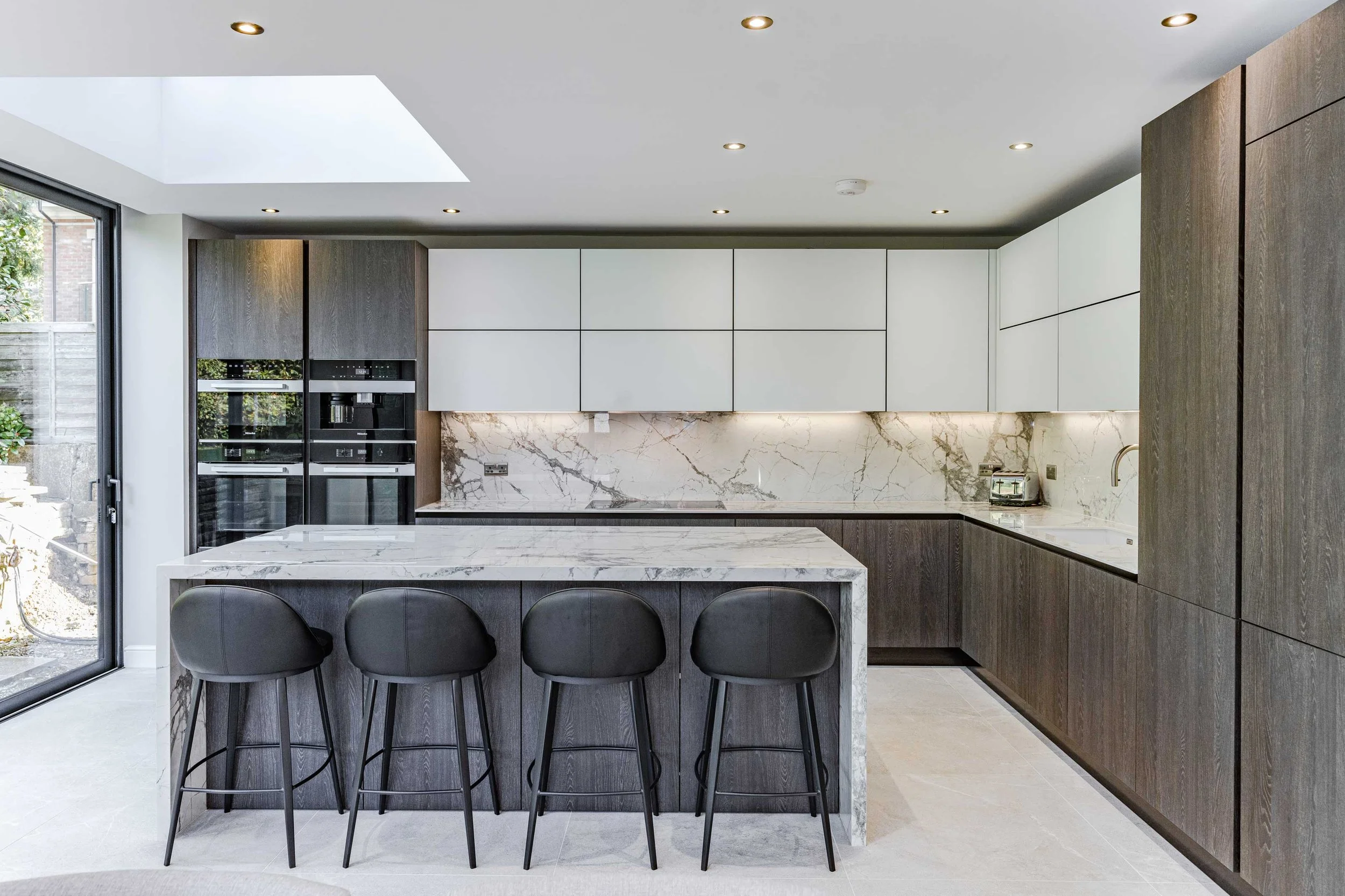 Modern kitchen with white and dark wood cabinets, a marble island with black bar stools, and a marble backsplash under ceiling lights, with a sliding glass door on the left.