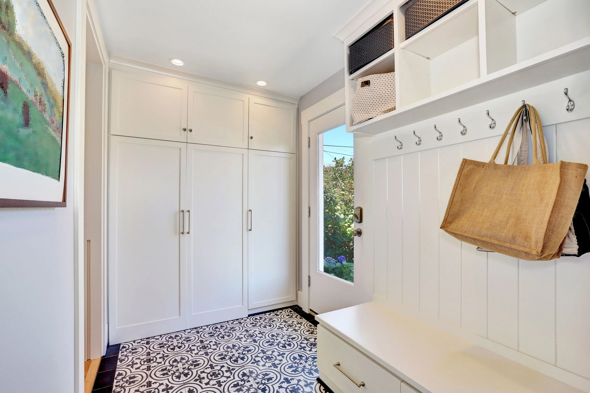 Entryway with white wooden wall paneling, hooks with a hanging tan bag, a built-in white bench, an upstairs storage with baskets, a window showing green foliage outside, and black and white patterned tile flooring.