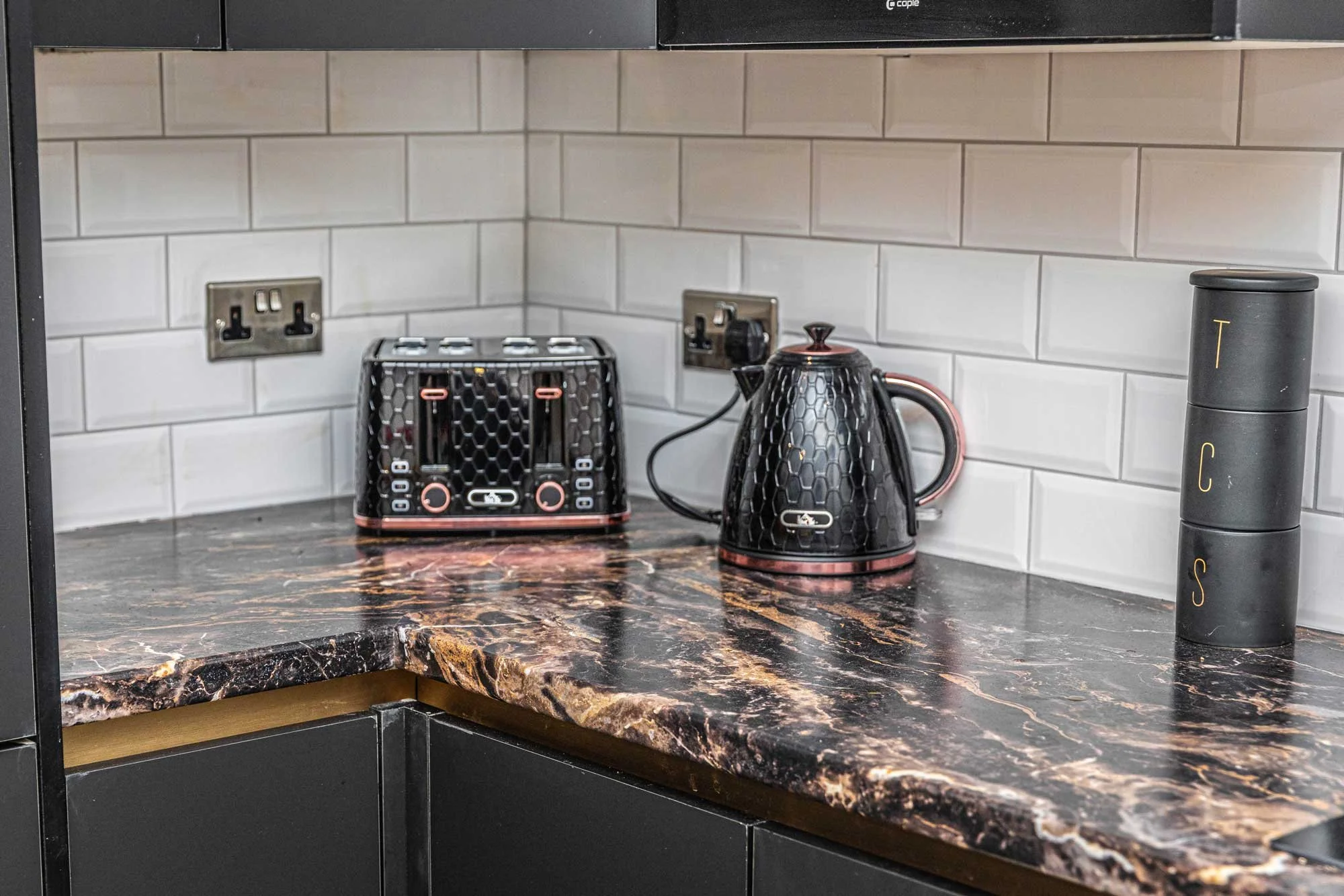 A kitchen countertop with a black honeycomb-patterned toaster and a matching electric kettle, both with copper accents. There's a white subway tile backsplash and a black appliance on the right side.
