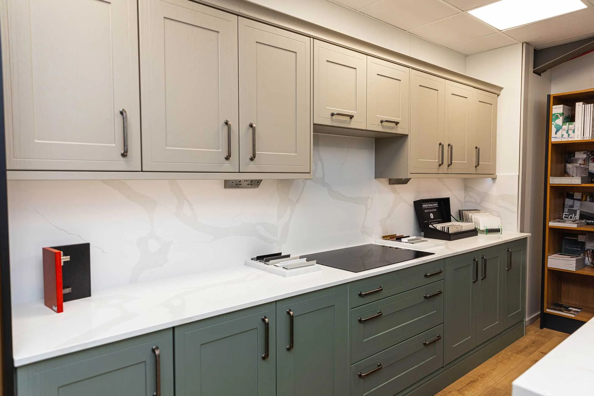 Showroom kitchen display with white upper cabinets, dark green lower cabinets, white marble backsplash, electric stove, and a bookshelf on the right side.