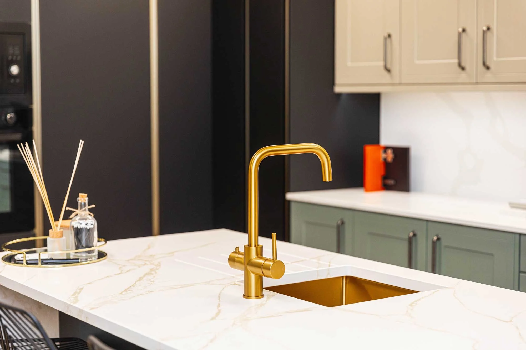 Modern kitchen with a white marble countertop, gold sink and faucet, black and beige cabinets, and kitchen accessories like reed diffusers and a tray.