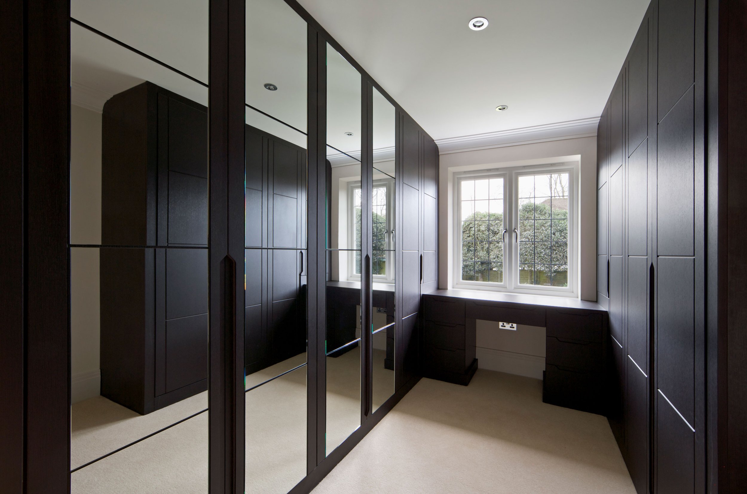 A walk-in closet with dark wood built-in cabinets and drawers, a large mirror, a window with a view of trees outdoors, and ceiling lights.