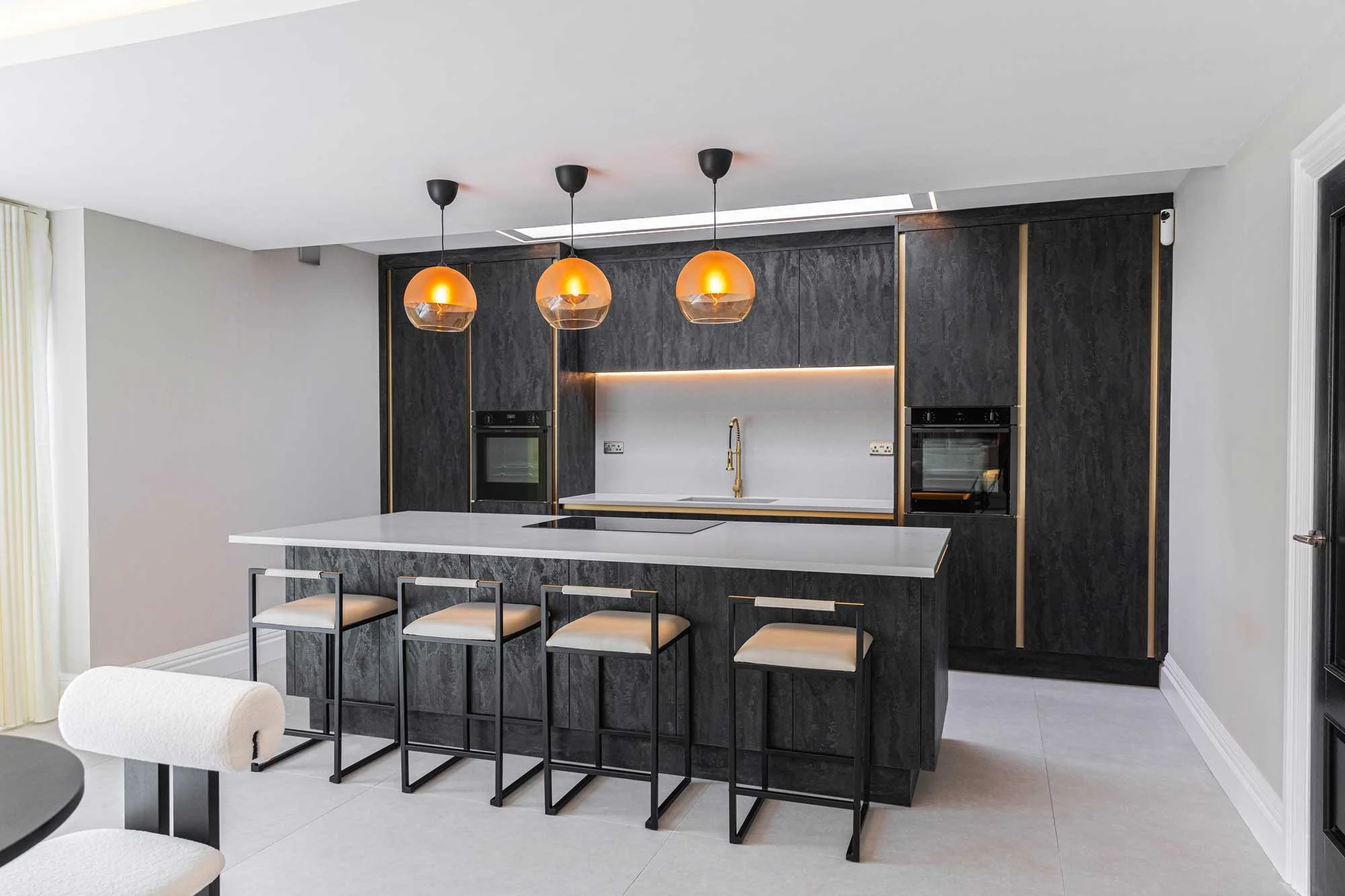 Modern kitchen with black cabinets, white countertop island, three pendant lights with orange shades, built-in oven, and bar stools.