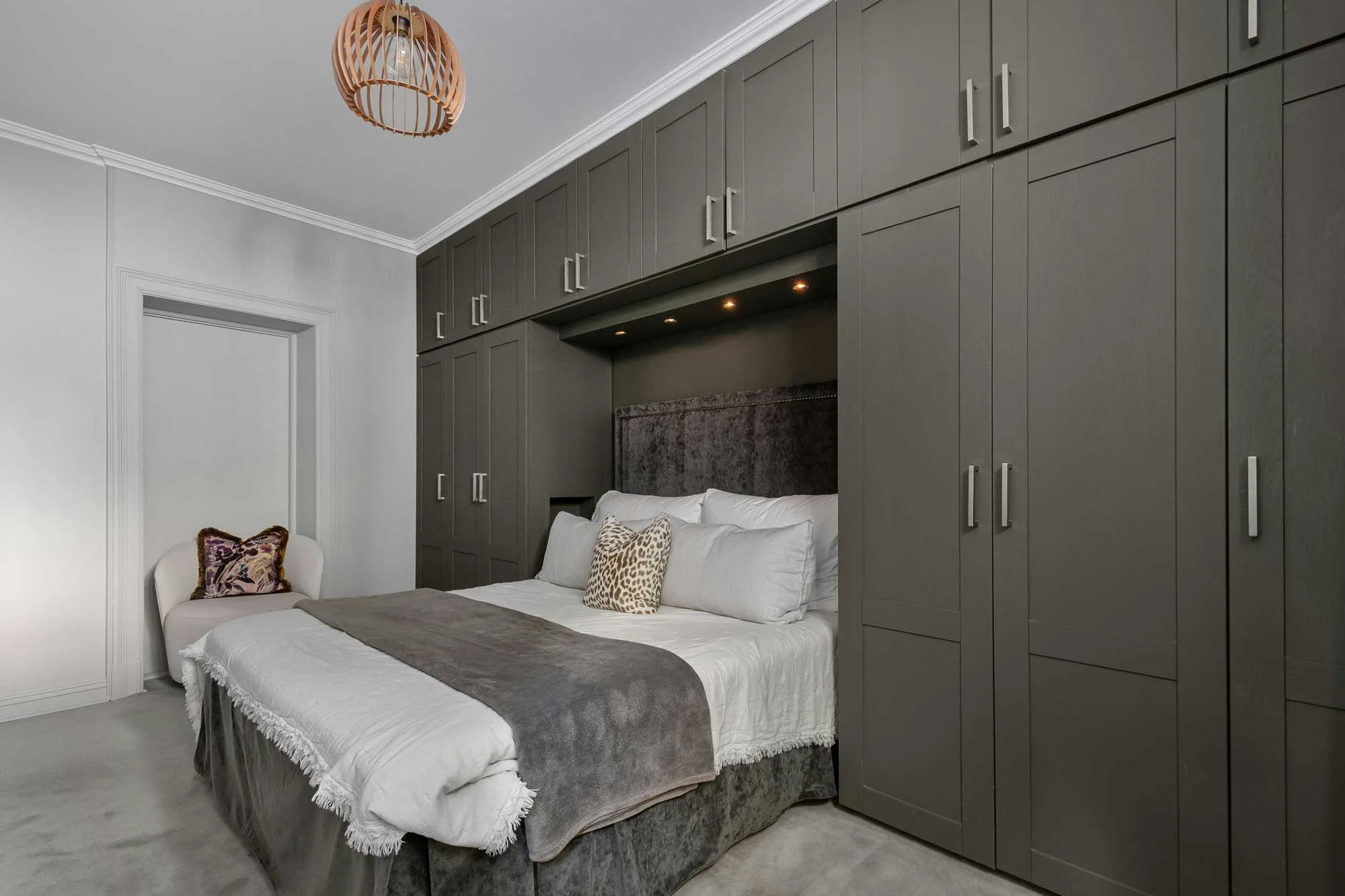 A bedroom with a bed against a dark gray built-in wall of cabinets, two decorative pillows, a white chair with a floral pillow in the corner, a gray throw blanket, a wooden pendant light fixture on the ceiling, and a white doorframe on the wall.