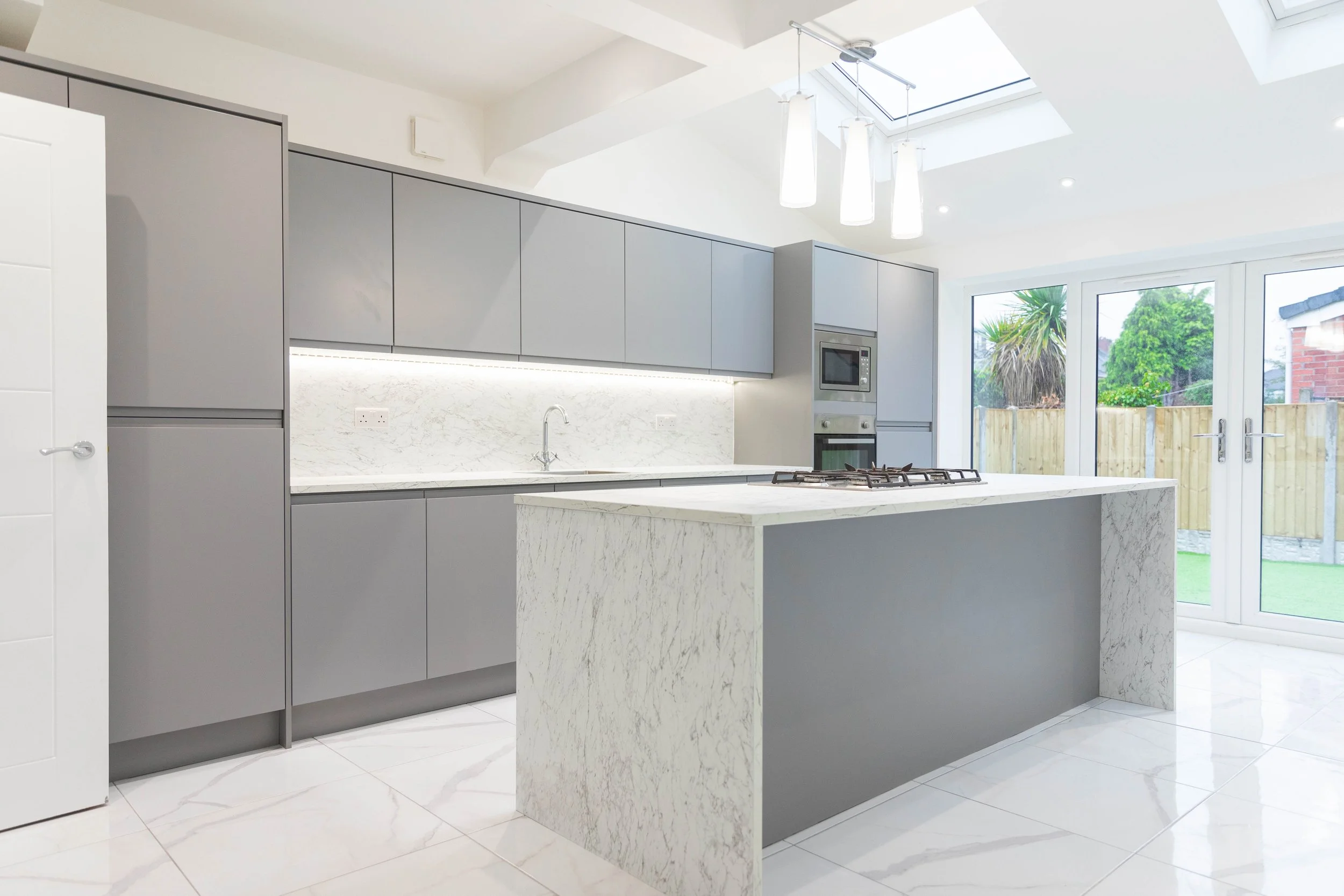 Modern kitchen with gray cabinets, white marble island, built-in microwave and oven, large glass doors leading outside, and skylights in a bright, minimalistic space.