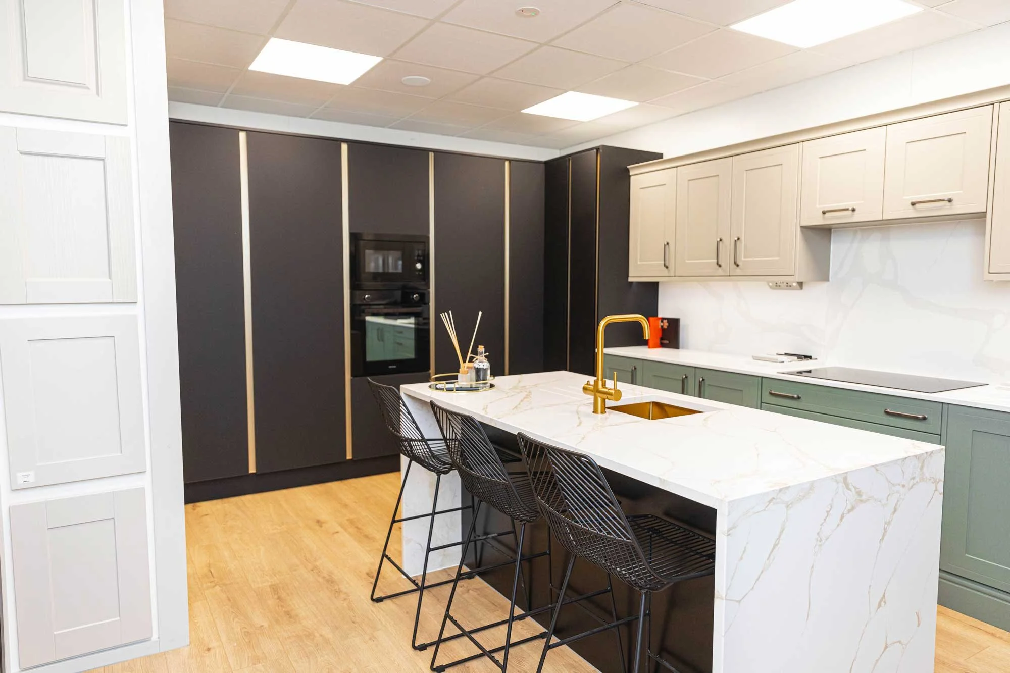 Modern kitchen with beige upper cabinets, dark black lower cabinets, and a white marble island with a gold faucet and black wire chairs.